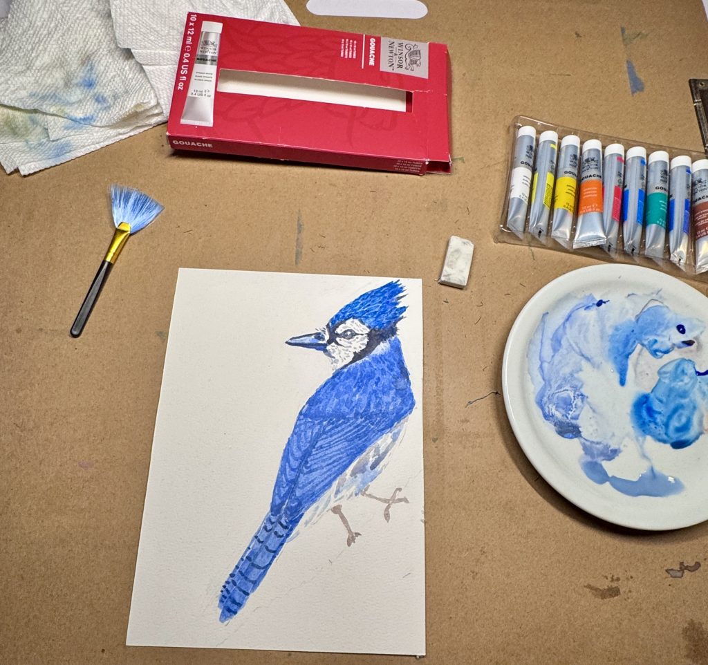 Blue jay gouache painting on a worktable beside Winsor & Newton paints, a brush & a plate used for mixing blue paint.